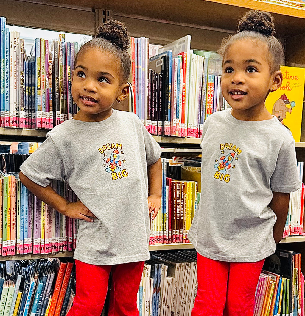 Two children wearing gray t-shirts with a colorful design in a library setting.